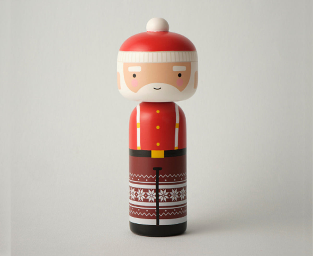 A Santa Claus-themed wooden kokeshi doll with a red hat, white beard, red jacket with yellow buttons, and a brown patterned skirt with white snowflakes, isolated on a gray background.