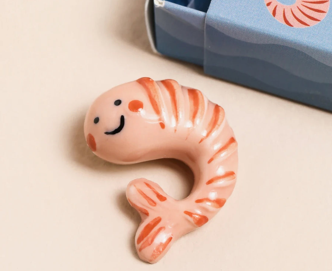 A pink and orange striped shrimp-shaped ceramic object with a smiling face, shown next to a blue box on a beige background.