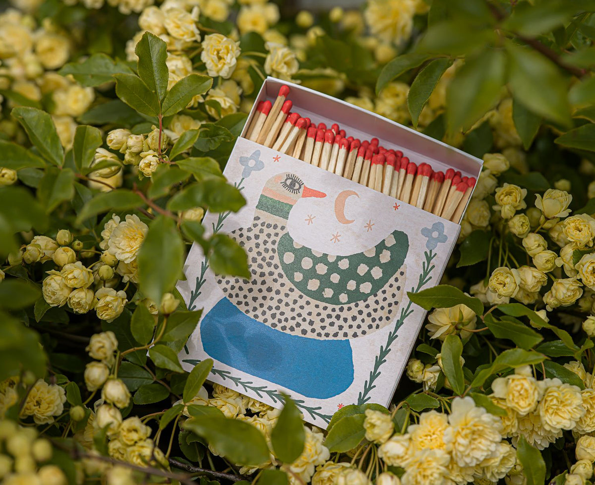 A decorative matchbox filled with wooden matches topped with red tips on a bed of flowers. The matchbox lid features a stylized spotted duck illustration in green, blue, black, and cream.