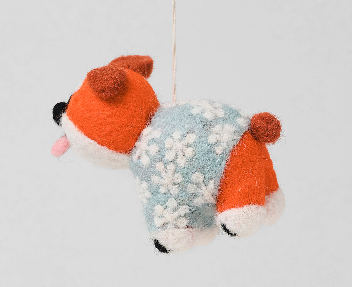 A handcrafted felt dog ornament wearing a light blue sweater with white snowflakes, hanging against a plain light gray background.