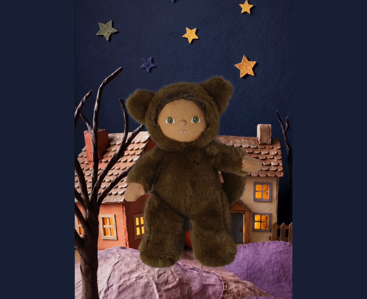 A plush werewolf cozy dinkum doll wearing a brown hooded outfit with floppy ears, posed in front of a handmade paper-cut village scene with houses, bare trees, and star decorations against a dark blue background.